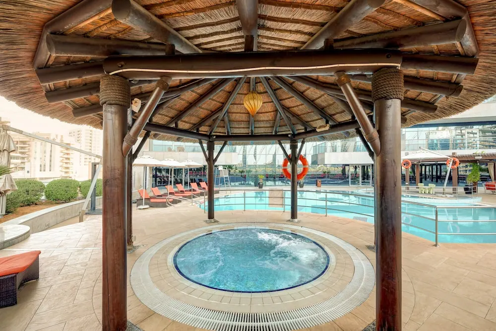 A thatched gazebo shelters a circular hot tub beside a large outdoor pool with orange sun loungers and umbrellas.