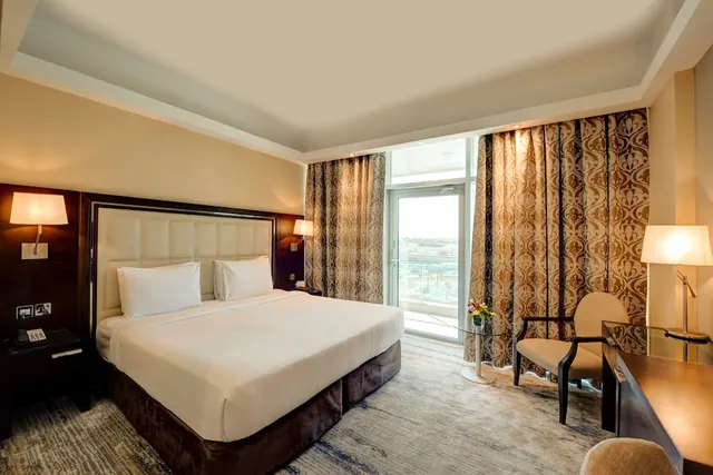 A hotel bedroom with a king bed, upholstered headboard, armchair, wooden desk, and a glass door leading to a balcony between patterned curtains.