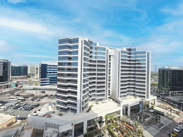 Elevated view of a white and glass high-rise building with a car park, street and outdoor seating area below under a clear blue sky.