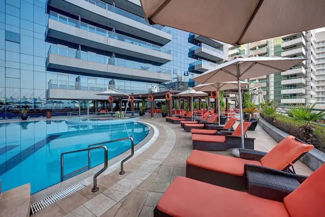 A curved swimming pool with orange sun loungers and shade umbrellas on the pool deck, with tall residential buildings behind.