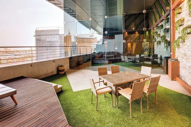 A covered rooftop terrace with artificial grass, a wicker dining set, a timber deck and a glass balustrade overlooking city buildings and cranes.