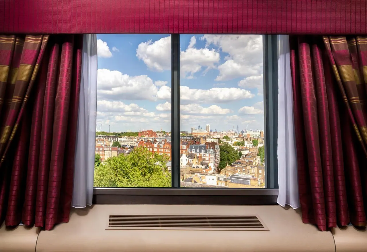 A window with burgundy curtains looks out over a city of rooftops, trees and brick buildings under a partly cloudy sky.