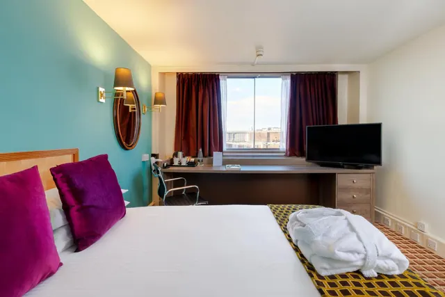 A hotel bedroom with a made double bed, a desk with a kettle and tea tray, a wall-mounted television, and a window showing distant buildings.