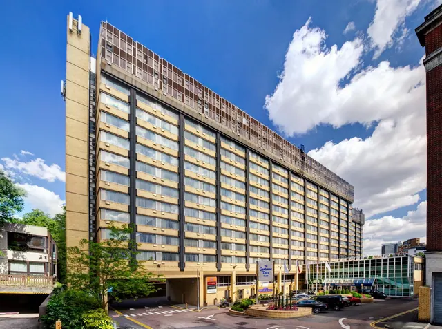 Exterior of a tall tan-brick hotel building with a forecourt, parked cars and a blue sky with clouds.