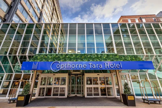 The front entrance of the Copthorne Tara Hotel, with a blue sign under a green wall, a glass facade, revolving doors and two luggage trolleys on the pavement.