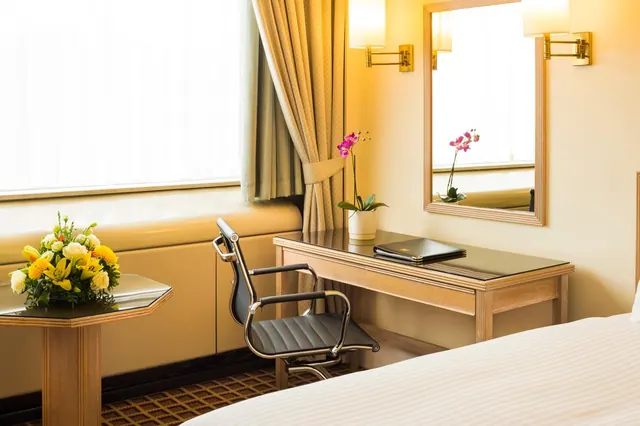 A hotel bedroom with a wooden desk, a black desk chair, a wall mirror, wall-mounted lamps, and a flower arrangement on a side table beside a bright window with gold curtains.