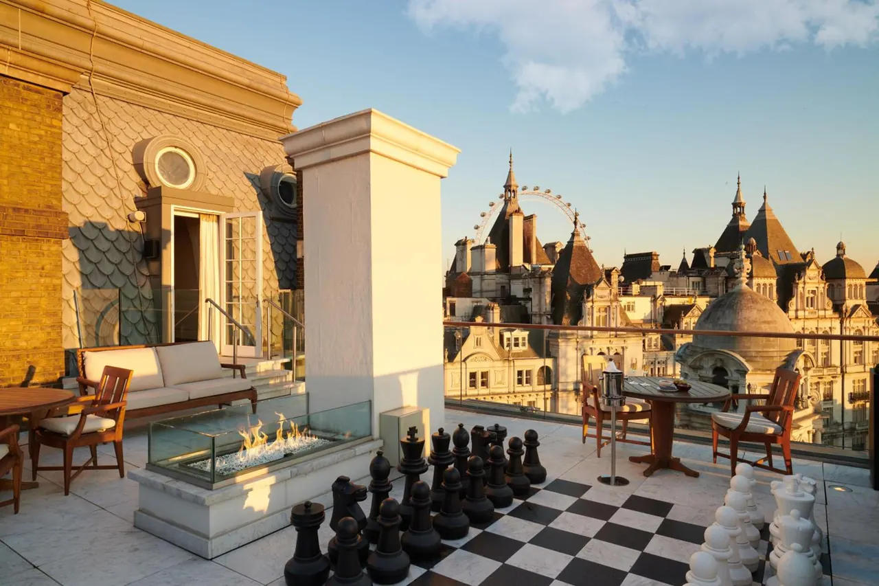 A rooftop terrace with a sofa, chairs, a gas fire pit and a large outdoor chessboard, with the London Eye and historic buildings visible beyond the railing.