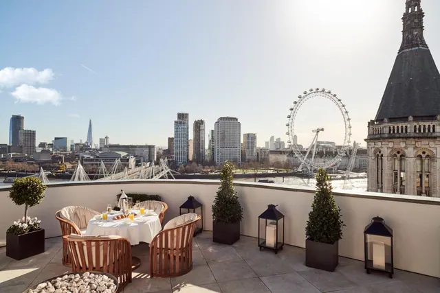 A rooftop terrace with a set breakfast table, wooden chairs, potted topiary and lanterns, overlooking the London Eye, city skyline and River Thames.