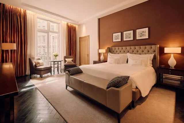 A bedroom with a king bed, tufted upholstered headboard, two armchairs and tall windows opening onto a balcony.