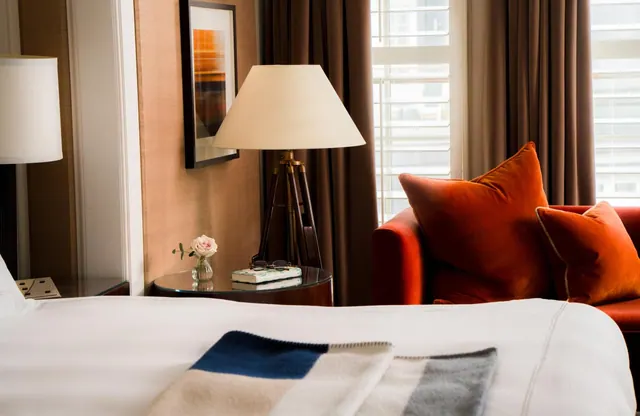 A hotel bedroom with a white bed and striped throw, an orange armchair with cushions, a tripod floor lamp and a side table holding a rose in a glass vase.