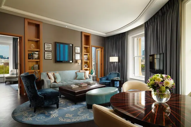 A living area with a blue sofa, armchairs, a round dark wood table with a flower arrangement, and wall-mounted televisions, with windows showing buildings outside.