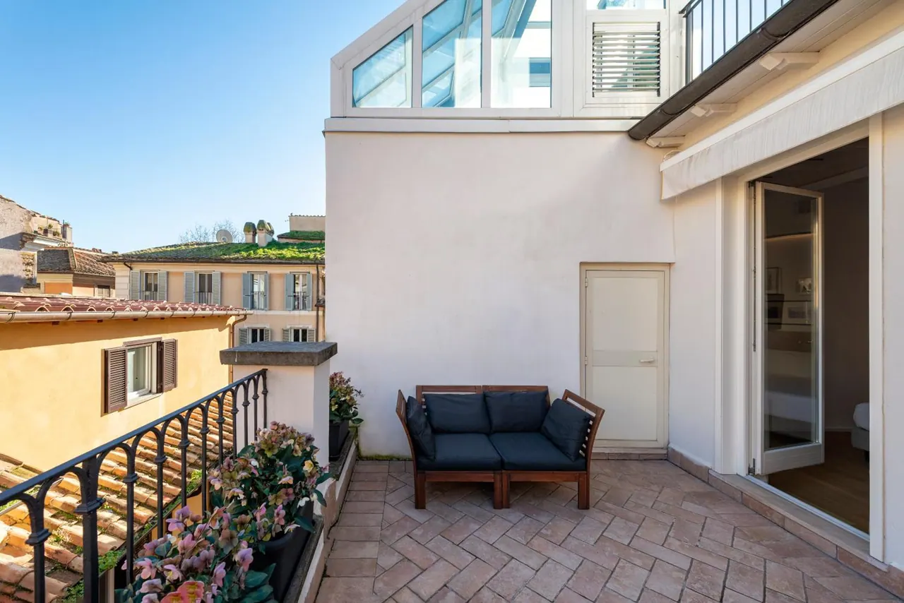 A brick-paved terrace holds a wooden sofa with navy cushions and potted flowers beside a metal railing, with rooftops and buildings beyond and an open door into a bedroom.