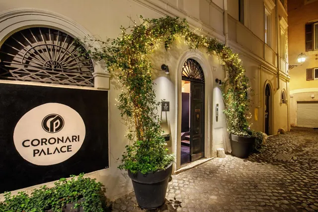 The lit entrance of Coronari Palace at night, with an open arched doorway, climbing plants strung with fairy lights, and a cobbled street in front.