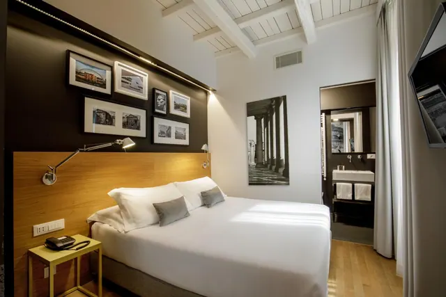 A double bed with white linen and grey cushions sits beside a dark wall of framed photos, with a wall-mounted television at right and an open bathroom doorway beyond.