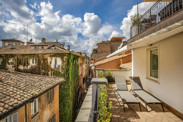 A tiled terrace with two grey sun loungers and potted plants overlooks narrow lanes, rooftops and an ivy-covered building under a cloudy sky.