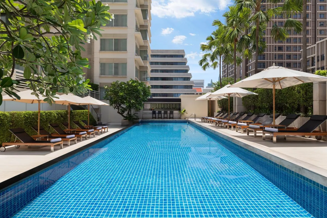 A rectangular blue-tiled pool with white parasols and dark sun loungers along its edge, with palm trees and high-rise buildings behind.