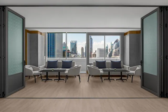 A lounge area with grey armchairs, sofas and low dark tables sits between folding glass doors, with windows showing city skyscrapers outside.