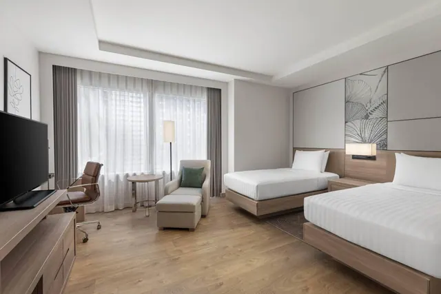 A hotel bedroom with two beds, a flat-screen television on a wooden console, a desk chair, an armchair with an ottoman, and a large window with sheer curtains.