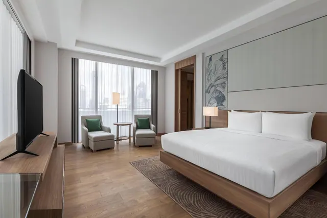 A bedroom with a king bed, two armchairs by a large window with sheer curtains that looks out toward city buildings, and a flat-screen television on a wooden stand.