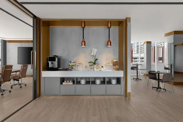 A breakfast station with a coffee machine and pastries sits against a grey wall, with bistro tables and city buildings visible through the windows beside it.