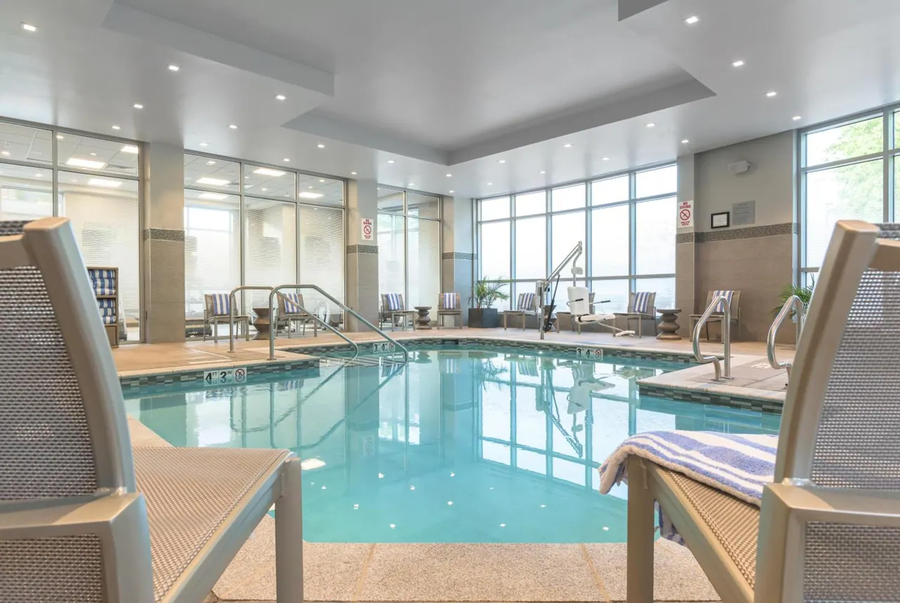 An indoor swimming pool with metal grab rails, a pool lift chair, lounge chairs and tall windows along one side.