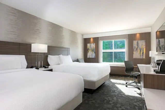 A hotel bedroom with two made beds, a window with green trees outside, and a desk with a chair on the right.