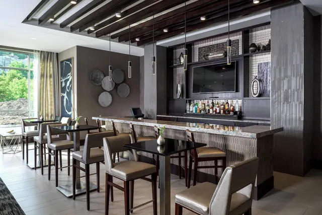 A bar area with high tables, bar stools, a bottle display behind the counter and a wall-mounted television, with a large window on the left.