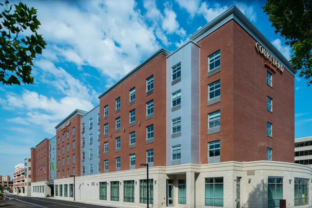 The exterior of a multi-storey brick hotel building with grey panelled sections and a stone-clad ground floor, under a partly cloudy blue sky, with a street and trees at the edges of the frame.