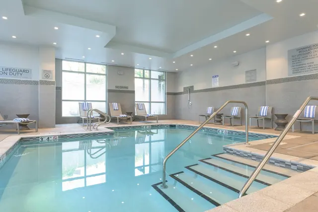 An indoor swimming pool with entry steps, metal handrails, and striped lounge chairs along the wall under recessed ceiling lights.