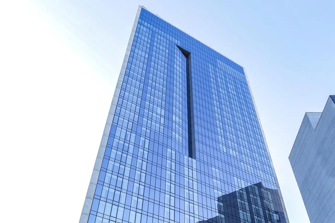 Low-angle view looking up at a tall blue glass tower with a vertical recessed slot against a pale sky, with part of another building at the right edge.