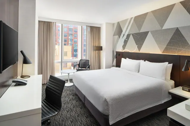 A bedroom with a king bed under white linens, a wall-mounted television, a desk with a chair, and a window looking out onto brick buildings.