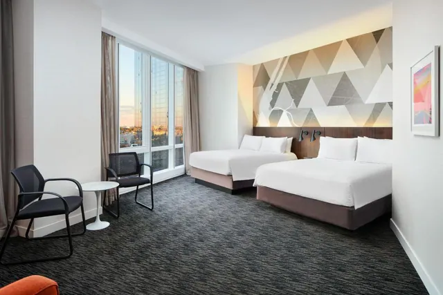 A hotel room with two made beds with white bedding, a geometric patterned feature wall, and floor-to-ceiling windows looking out on city buildings, with two armchairs and a small round table by the window.