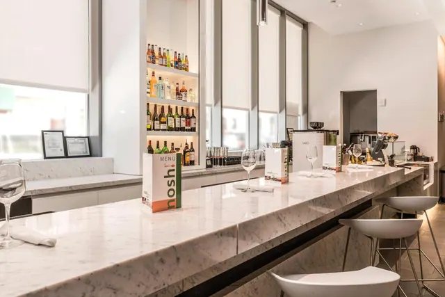 A long marble bar counter with white stools, wine glasses and menus sits in front of a shelf of bottles and tall windows with roller blinds.