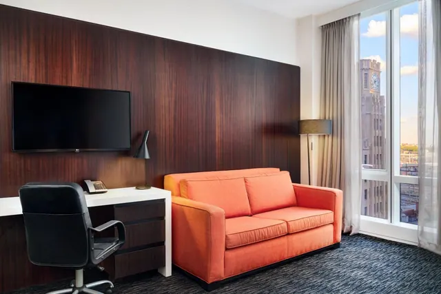 A hotel room with a wall-mounted television, a desk with a black chair, an orange sofa, and a large window looking out at a clock tower building.