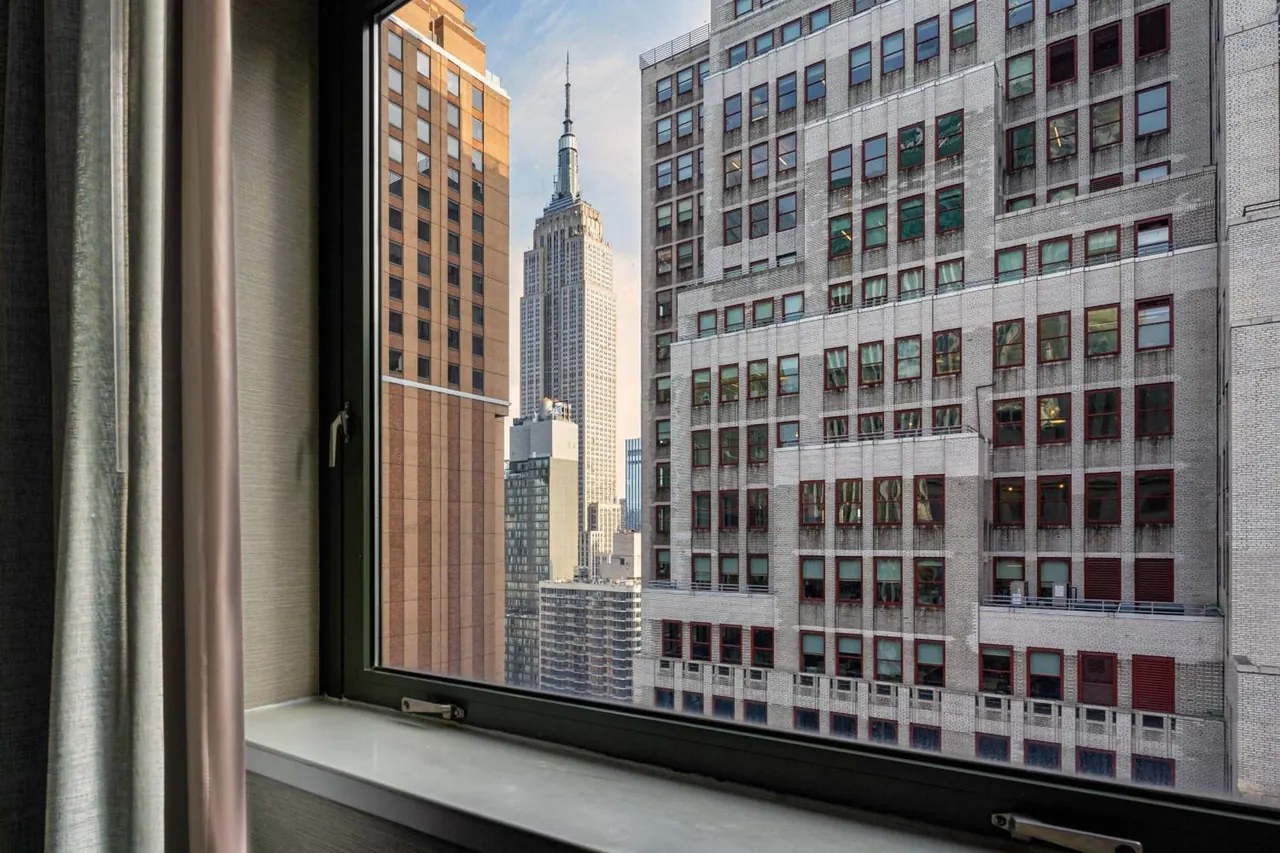 A window with a dark frame and light sill looks out over surrounding midtown towers to the Empire State Building, with curtains framing the left side.