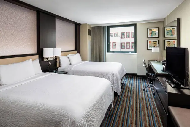 A hotel room with two beds made up with white bedding, a wall-mounted television, a desk with a chair, and a window facing a building across the street.