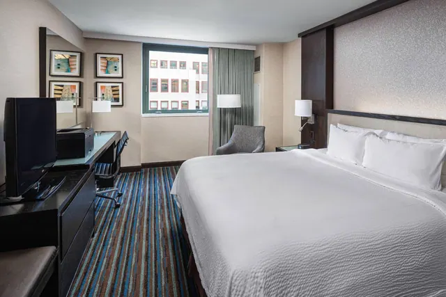 A king bed with white bedding faces a desk, television and armchair in a hotel room with a window onto a neighbouring building.