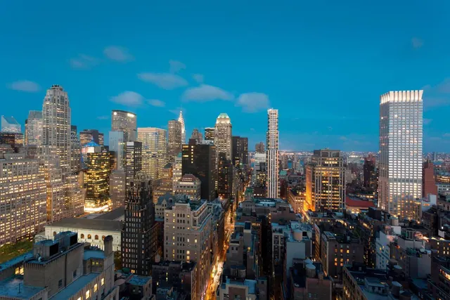 Aerial view over a dense Manhattan skyline at dusk, with illuminated office towers and the Chrysler Building spire visible.