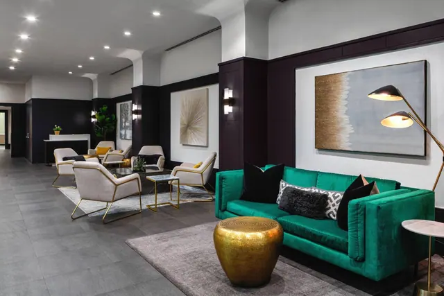 A lobby seating area with a green velvet sofa, cream armchairs with gold frames, a brass pouf, large abstract wall art and a floor lamp, with a reception desk visible in the background.