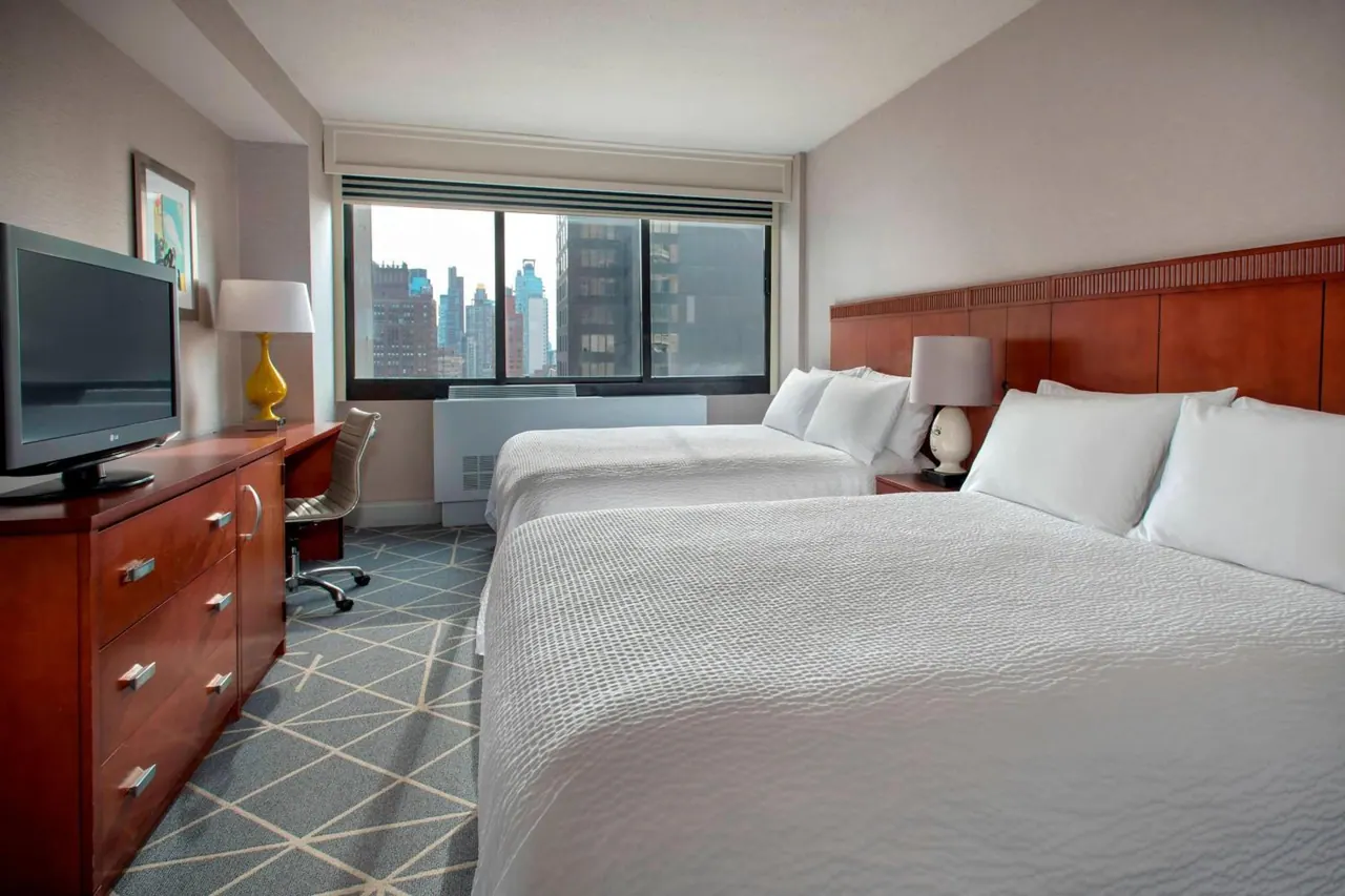 A hotel room with two beds made with white bedding, a flat-screen TV on a wooden dresser, a desk chair, and a window showing city skyscrapers.