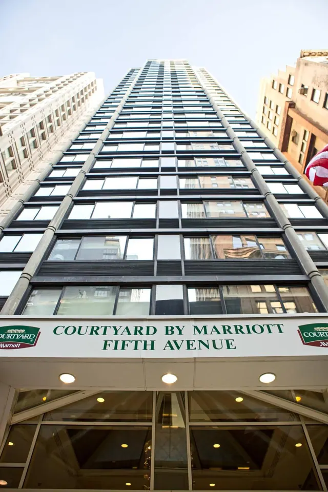 Looking up at a tall glass-and-steel hotel tower, with the Courtyard by Marriott Fifth Avenue sign above the entrance.