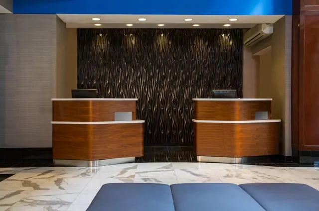 Hotel reception area with two wooden front desks, a textured dark wall panel, a marble floor, recessed ceiling lights and a wall-mounted air conditioner.