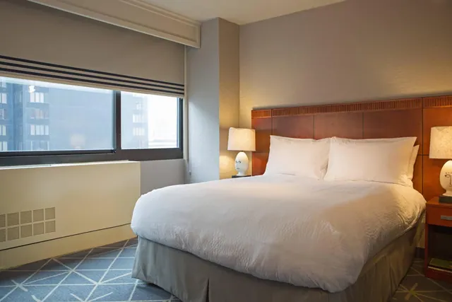 A made king bed with white linens and a wooden headboard sits between two bedside lamps, with a window beside it showing neighbouring buildings.