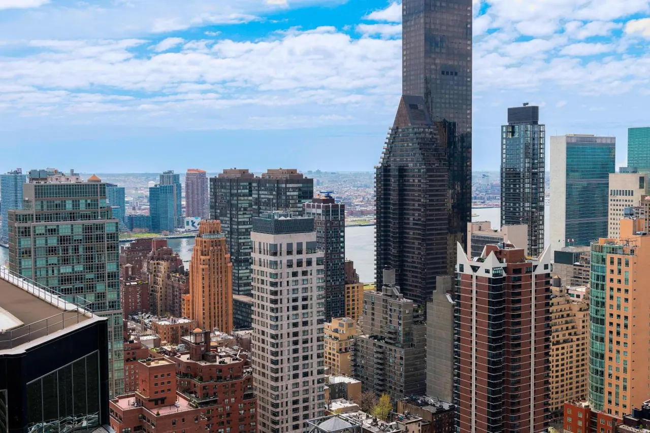 Elevated view over dense midtown Manhattan towers, with a river and distant low-rise buildings visible under a partly cloudy sky.