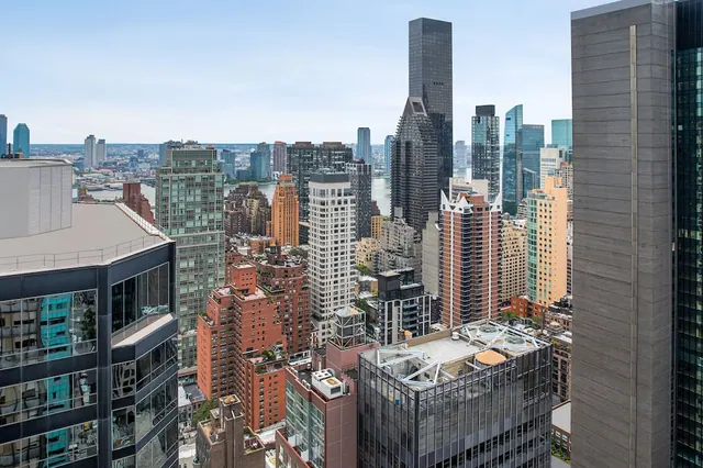 Elevated view over rooftops and high-rise towers toward a river and the distant Queens skyline under a pale sky.