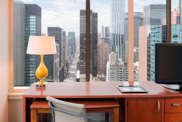 A wooden desk with a yellow table lamp, a flat-screen television and a white chair sits in front of large windows overlooking a city avenue lined with skyscrapers.