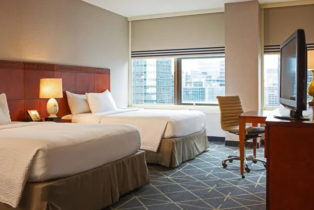A hotel bedroom with two double beds, a desk and chair, a flat-screen television, and a window looking out onto city buildings.
