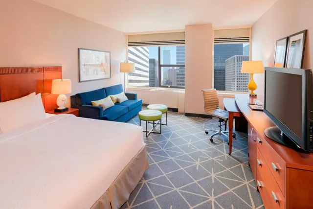 A hotel bedroom with a king bed, a blue sofa, a work desk and a television on a wooden dresser, with a window showing office towers outside.