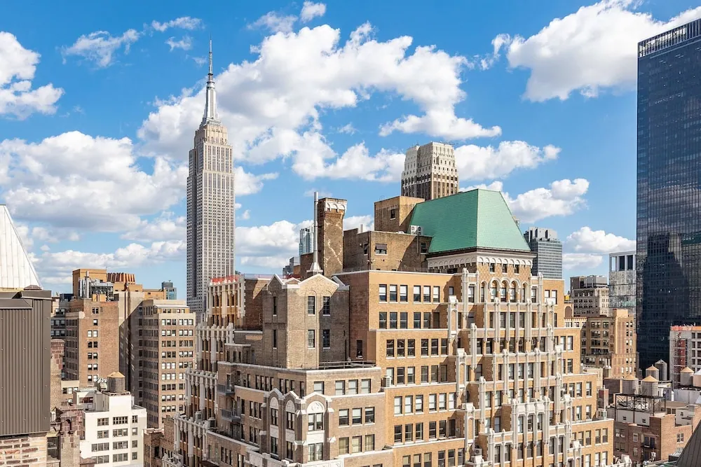 View across midtown Manhattan rooftops toward the Empire State Building under a partly cloudy blue sky.