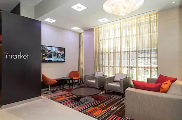 A lobby lounge with a grey sofa and armchairs, red and orange cushions, a wall-mounted television, and a dark panel sign reading 'the market'.